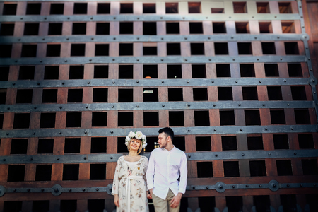 Portrait of wedding couple standing isolated on attractive architecture . They holding their hands, husband looking at his wife and she is smillingの写真素材