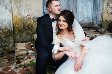 Fashionable wedding couple sitting near old house and smiling. Husband holding his wife. Bride wearing bridal veil and dress with embroidery.の写真素材