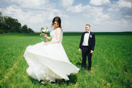Beautiful wedding couple walking in the green field. Bride carrying her wedding bouquet and spinning. Groom looking at his bride.の写真素材