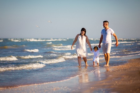 Young happy family walking along the seashore at the day time. Parents holding hands of their son.の写真素材