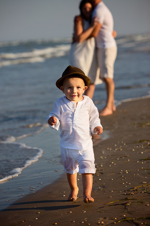 Young happy family walking along the seashore at the day time. Little baby boy walking in the foreground, his wearing a cool brown hat. Parents staying on the background and holding each other.の写真素材