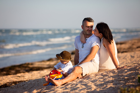 Young happy family sitting on the beach on the day time. Mother hug his husband behind his back, parents looking to each other and smiling. Little baby boy playing with the sand, his wearing a cool brown hat.の写真素材