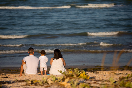 Young happy family sitting on the beach on the day time. They looking at the sea. Little baby boy playing with the sand, his wearing a cool brown hat.の写真素材