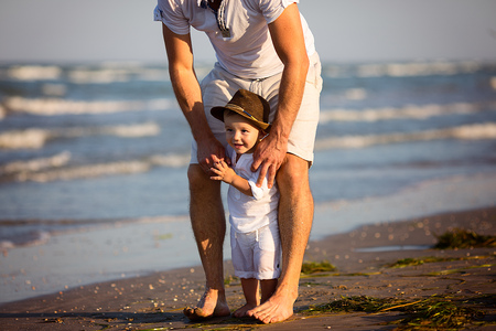 Young happy family walking along the seashore at the day time. Mother holding her daughter by the hand, looking at she and smiling. Little baby girl wearing beautiful white dress and a hat.のeditorial素材