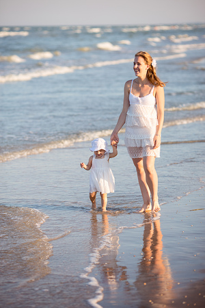 Young happy family walking along the seashore at the day time. Mother holding her daughter by the hand, looking forward and smiling. Little baby girl wearing beautiful white dress and a hat.の写真素材