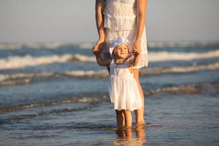Young happy family walking along the seashore at the day time. Mother holding her daughter by the hand, it is only her lower body on the photo. Little baby girl playing with water, she is wearing beautiful white dress and a hat.の写真素材