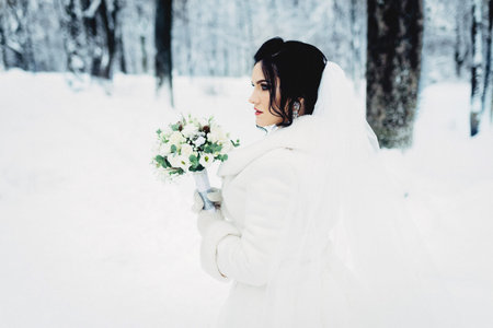 Portrait of an amazing bride. She walking in the winter forest full of snow, wearing white dress and fur coat, carrying her wedding bouquet.の写真素材