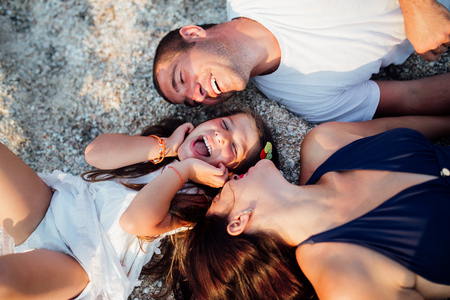 Young happy family walking along the seashore. Little girl with her parents lying on the sand and smiling, parents want to kiss their girl on the cheek.の写真素材