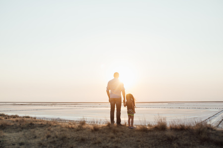 Young happy family walking along the seashore. Little girl running ahead and wearing white dress, parents walking behind her, embracing each other and smiling.の写真素材