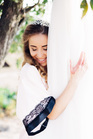 Nice portrait of a beautiful bride with a diadem on her head. She is standing in the yard under the tree, smiling and holding her wedding dress in her hands.の写真素材