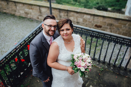 Portrait of a beautiful wedding couple. They are standing on a balcony of an old building, a groom embracing his bride and they are smiling and looking at the camera.の写真素材