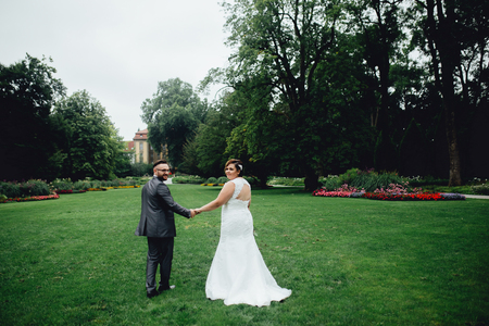 Portrait of a beautiful wedding couple. They are walking in the park, between a big green trees, holding their hands. Looking back at the camera.の写真素材