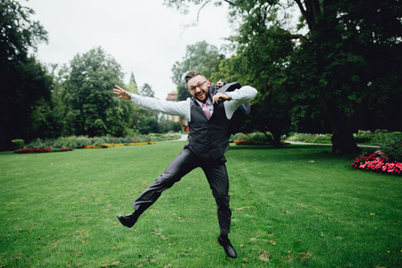 Portrait of a handsome groom in a grey suit with a tie and vest. He is  jumping in the park, on a green grass and near a big trees, holding his jacket in one hand, looking at the camera and smiling.の写真素材