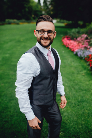 Portrait of a handsome groom in a grey suit with a tie and vest. He is walking in the park, on a green grass and near a big trees. Looking at the camera and smiling. He is wearing eyeglasses.の写真素材