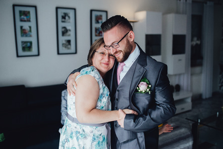 Portrait of a handsome groom in a grey suit with a tie with his mother. They are standing in the dining room, embracing each other and smiling.の写真素材