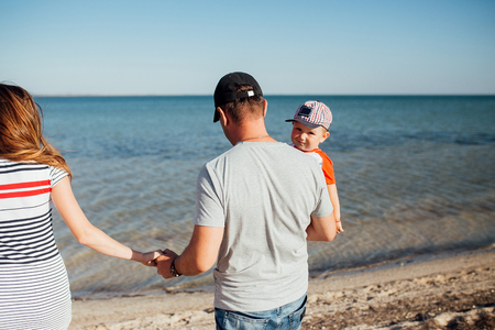 Funny portrait of a happy family on the beach. They are walking on the beach. Father is holding his son on his hands and his wife by the hand. They standing back to the camera. A boy looking at the camera.の写真素材