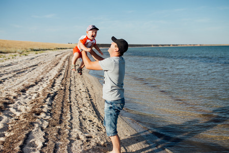 Funny portrait of a father and son, while they are playing on the beach. Father is holding his boy on his hands  and throwing him up. Little boy is flying and smiling.の写真素材