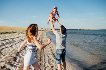 Funny portrait of a happy family on the beach. They are walking on the beach. Father is holding his son on his hands and throwing him up, mother is running behind them.の写真素材