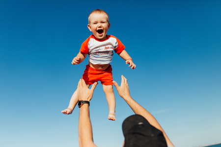 Funny portrait of a father and son, while they are playing on the beach. Father is holding his boy on his hands  and throwing him up. Little boy is flying and smiling.の写真素材