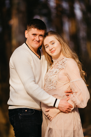 Beautiful portrait of a pregnant woman and her husband. They are standing face to face near the tree, embracing and looking at the camera.の写真素材