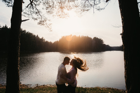 Beautiful portrait of a pregnant woman and her husband. They are standing face to face near the river, embracing, during sunset.の写真素材