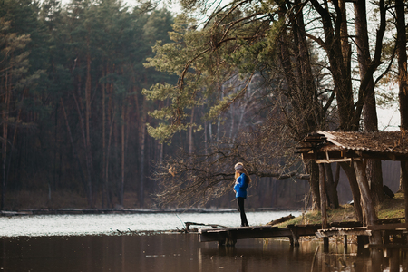 Beautiful portrait of a pregnant woman in the forest. She is standing near the river, holding her tummy and looking at it.の写真素材