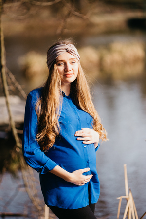 Beautiful portrait of a pregnant woman in the forest. She is standing near the river, holding her tummy and looking away.の写真素材