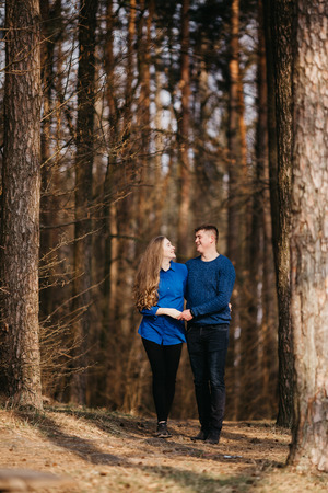 Beautiful portrait of a pregnant woman and her husband. They are walking in the forest, holding their hands and looking at each other. They are smiling.の写真素材