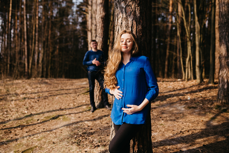 Beautiful portrait of a pregnant woman and her husband. They are walking in the forest, standing far from each other near the trees and smiling.の写真素材