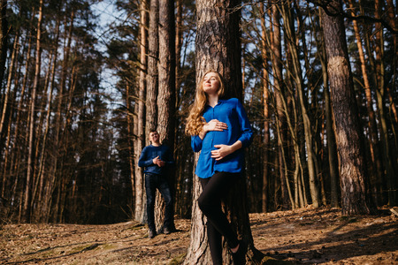 Beautiful portrait of a pregnant woman and her husband. They are walking in the forest, standing far from each other near the trees and smiling.の写真素材