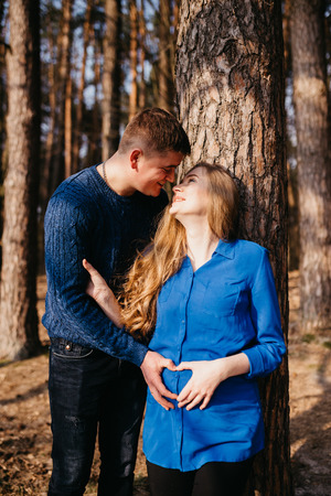 Beautiful portrait of a pregnant woman and her husband. They are walking in the forest, standing near the tree and embracing.の写真素材
