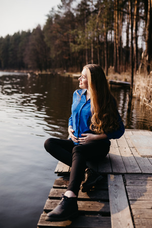 Beautiful portrait of a pregnant woman in the forest. She is sitting near the river, holding her tummy and looking away.の写真素材