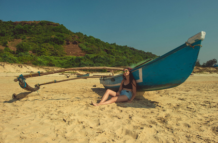 Girl sitting on the sand near the blue boat against the background of the hillの写真素材