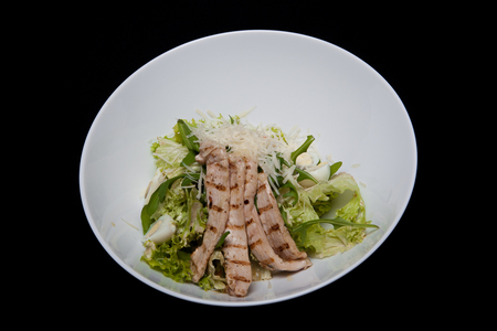 Close-up of tasty fresh caesar salad with chicken and vegetables decorated with balsamic sauce on white plate rim on black table.の写真素材