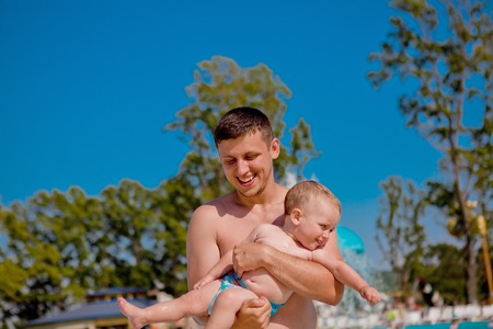Young active father playing with a cute baby, happy laughing boy, enjoying a hot summer day in an outdoor pool of a resort during vacation.の写真素材