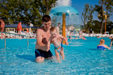 Young active father playing with a cute baby, happy laughing boy, enjoying a hot summer day in an outdoor pool of a resort during vacationの写真素材