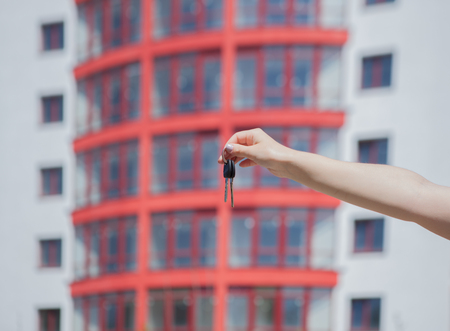 Female hand holding keys on the background of new house. Real estate concept. Keys to new apartment. Moving home or renting property. Real estate agent.の写真素材