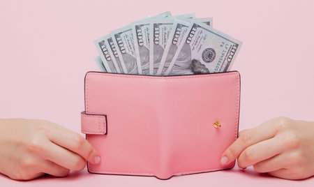 Dollars and pink wallet in woman's hand on a pink background with copy space.