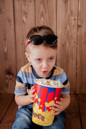 Little cute kid baby boy 2-3 years old , 3d cinema glasses holding bucket for popcorn, eating fast food on wooden background. Kids childhood lifestyle concept. Copy space.の写真素材
