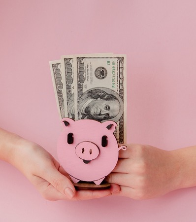 Women's hands holding pink piggybank and dollar banknotes on pink background.の写真素材