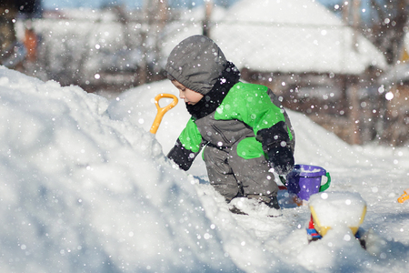Portrait of cute little toddler sitting on snow and playing with his yellow tractor toy in the park. Child playing outdoors. Happy boy with construction toy. Lifestyle concept.の写真素材