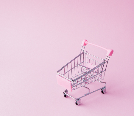 Small supermarket grocery push cart for shopping toy with wheels and pink plastic elements on pink pastel color paper flat lay background. Concept of shopping. Copy space for advertisement.の写真素材
