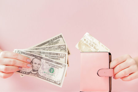 Dollars and pink wallet with credit card in woman's hands on pink background.
