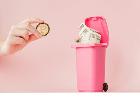 bitcoin in a woman's hand and dollars in a trash can on pink background.