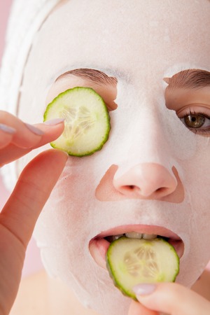 Beautiful young woman is applying a cosmetic tissue mask on a face with cucumber on a pink background.の写真素材