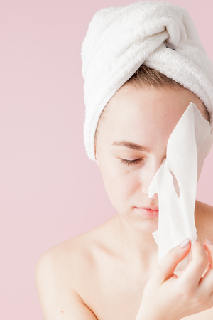 Beautiful young woman is applying a cosmetic tissue mask on a face on a pink background. Healthcare and beauty treatment and technology concept.の写真素材