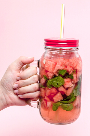Young woman hands holding freshly squeezed watermelon lemonade of citrus fruits. Female with mason jar full of cold cocktail, mint leaves. Pink background, copy space, close up.の写真素材