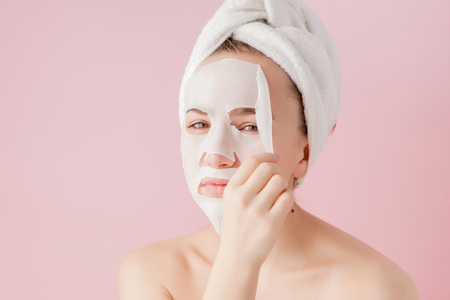Beautiful young woman is applying a cosmetic tissue mask on a face on a pink background. Healthcare and beauty treatment and technology concept.の写真素材