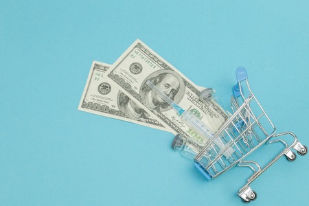 Medical injection and dollars in shopping trolley on blue background. Creative idea for health care cost, drugstore, health insurance and pharmaceutical company business concept. Copy space.の写真素材