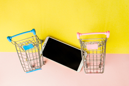 Blank screen white cellphone and shopping carts on pastel pink and yellow background. Minimal style, flatlay.の写真素材
